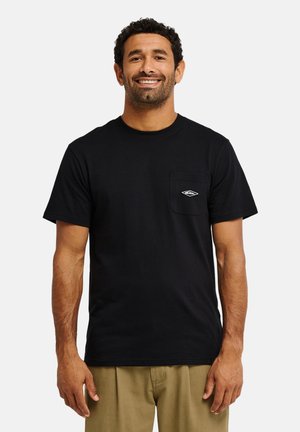 Homme aux cheveux courts et bouclés, portant un T-shirt noir à manches courtes avec un petit logo sur la poche poitrine et un pantalon beige, souriant.