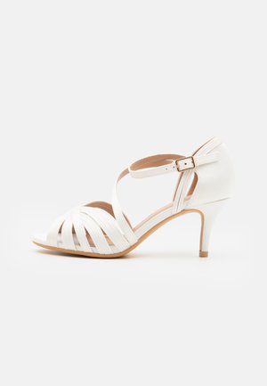 Valge kontsaga sandaal, millel on rihmaga disain. Omab peent pahkluu rihma ja klambrit ning siledat, tekstureeritud viimistlust peal.