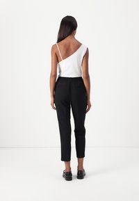 Hvit asymmetrisk tanktop med tynn stropp, kombinert med svarte taperede bukser med elastisk midjebånd, vist fra baksiden.