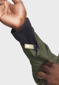 Grøn jakke med en mørk fleece indvendig manschet, en tan-farvet detalje med trykknap og et tætsiddende ærmedesign.