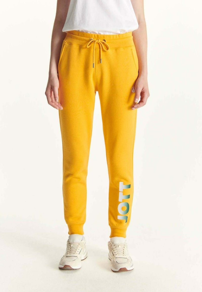 Geel joggingbroek met een elastische tailleband en trekkoord. Voorzien van een witte en teal grafische tekst "JOTT" op het linkerpootje. Zachte stof.