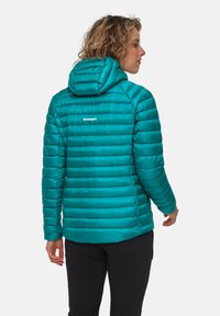 Kvinna som vänder ryggen mot kameran, iklädd en teal-färgad quiltad Mammut-hoodie och svarta byxor mot en neutral bakgrund.