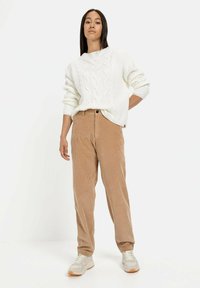 Cremefarvet kabelstrikket sweater med høj hals, parret med lysebrune fløjlshorts. Modellen har beige sneakers på og står imod en hvid baggrund.