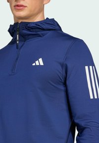 Plavi atletski pulover s pola-zip dizajnom, laganom teksturom i kapuljačom. Ima bijeli logo i tri vertikalne pruge na rukavu.