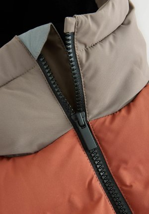 Steppjacke mit einem zweiseitigen Design: dunkelbrauner oberer Teil und rostfarbener unterer Teil, ausgestattet mit einem schwarzen Reißverschluss und einer weichen, glatten Textur.
