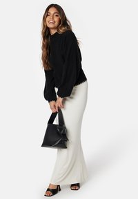 Maglione nero a trama con maniche a sbuffo, abbinato a una gonna lunga bianca e una borsa nera con dettaglio a fiocco. Sandali completano l'outfit.