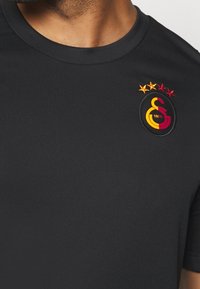 Närbild av en person som bär en svart tröja med rund hals och en röd- och guldfärgad sportlagssymbol med fyra stjärnor på bröstet.