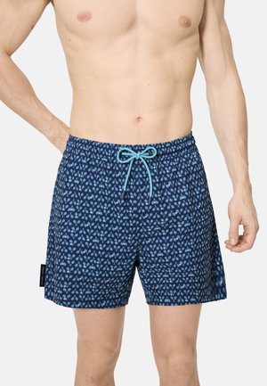 Hombre con pantalones cortos de baño azul marino con patrón de raquetas de tenis en azul claro y cintura con cordón, de pie contra un fondo liso.
