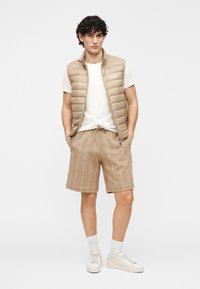 Smėlio spalvos pūkinė liemenė ant baltos marškinėlių, derinama su smėlio spalvos šortais su raštuotu dizainu. Lengvi sportbačiai ir balti kojinės užbaigia įvaizdį.