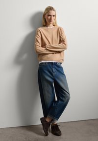 Maglione beige chiaro con texture a coste, abbinato a jeans in denim blu a vita alta e sneaker scamosciate marrone scuro.