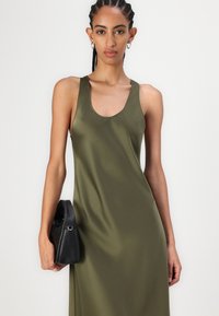 Rochie fără mâneci de culoare verde măsliniu cu decolteu rotund, realizată dintr-un material neted. Modelul ține în mână o poșetă mică neagră, cu un design elegant.