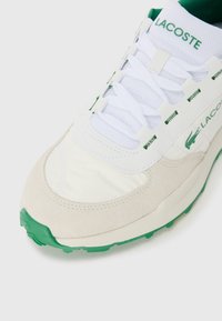 Witte en beige sneaker met groene accenten, van gestructureerd suède en mesh materialen, voorzien van veters en een logo op de zijkant.