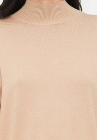 Μπεζ ριγωτό πουλόβερ με ζιβάγκο, εφαρμοστό στη λαιμόκοψη, με απαλή υφή και άνετη, χωρίς ραφές κατασκευή. Μια ελαφριά πλεκτή μπλούζα κατάλληλη για στρώσεις ρουχισμού.