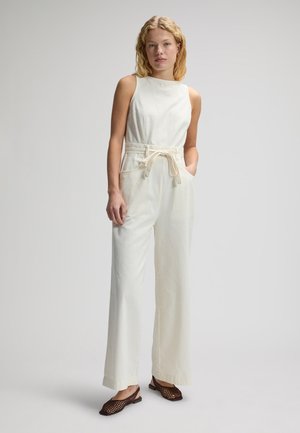 Kvinde stående iført ærmeløs cremefarvet jumpsuit med bindebælte og brune flettede flade sko, neutral baggrund.