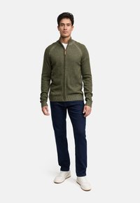 Cardigan zippé vert en tricot avec une finition texturée, présentant des manches raglan et une tirette en bois, associé à un jean en denim foncé et des baskets blanches.