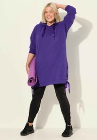 Fialová oversized mikina s tkaničkami, bočním šněrováním, zkombinovaná s černými legínami a černými teniskami. Model drží podložku na jógu.