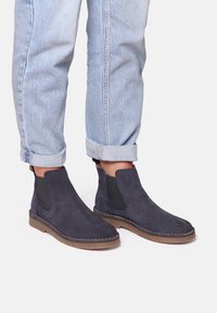 Marineblaue Wildleder-Stiefeletten mit elastischen Seiteneinsätzen, genähten Details und Gummisohlen, kombiniert mit hochgekrempelten hellblauen Jeans.