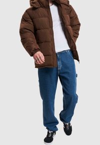 Brun vadderad jacka med huva, vit t-shirt under, blå baggy jeans och svarta sneakers. Inga synliga mönster eller logotyper.