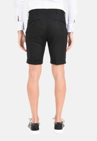 Pantaloni corti neri con orlo arrotolato, due tasche posteriori con bottoni. Indossati con sneaker nere con suole bianche. Tessuto dalla texture liscia.