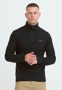 Sort zip-up sweatshirt med et tekstureret strikdesign, der har en høj krave og ribbede manchetter. En diskret logo-patch på brystet tilføjer detalje.