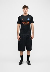 Νέος άνδρας στέκεται μπροστά σε λευκό φόντο φορώντας μαύρο αθλητικό μπλουζάκι με λογότυπα, μαύρο μακρύ σορτς, μαύρες κάλτσες και μαύρα παπούτσια.