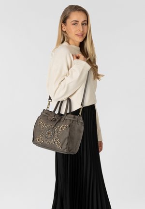 Donna con un maglione crema e una gonna plissettata nera tiene sulla spalla una borsa grigia decorata con borchie dorate e d'argento.