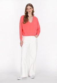Felpa corallo con scollo a V e maniche lunghe, vestibilità morbida. Abbinata a jeans bianchi a gamba larga e sneakers bianche, il tutto su uno sfondo neutro.
