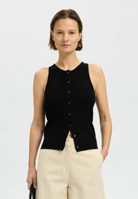 Top in maglia nero senza maniche con texture a coste, scollatura rotonda e chiusura frontale con bottoni; abbinato a pantaloni larghi di colore chiaro.