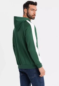 Felpa verde con strisce bianche ai lati; dotata di cappuccio regolabile con lacci, polsini e orlo a coste. Il tessuto ha una consistenza liscia.