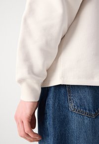 Pamut keverék pulóver puha fehér színben, bordás mandzsettákkal; párosítva kék farmernadrággal, klasszikus öt zsebes kialakítással.