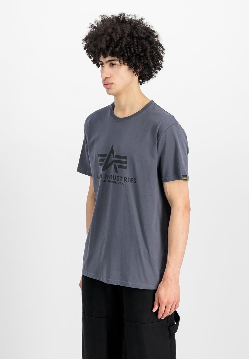 Graues Baumwoll-T-Shirt mit kurzen Ärmeln, das in der Mitte ein schwarzes Druckdesign und ein kleines Logo-Tag am linken Ärmel hat.
