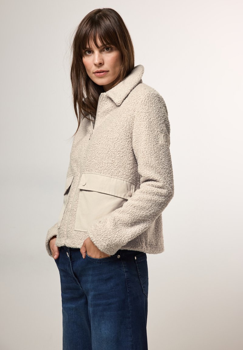Beige fleece jack met textuur, een kraag, een rits aan de voorkant, twee lichtgekleurde zakken en een logopatch op de linkermouw. Model draagt donkere jeans.