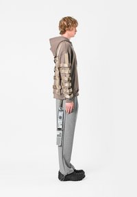 Sudadera con capucha en beige con acentos tejidos, combinada con pantalones de chándal grises y zapatos negros de tejido texturizado sin cordones. Etiqueta colgada de los pantalones.