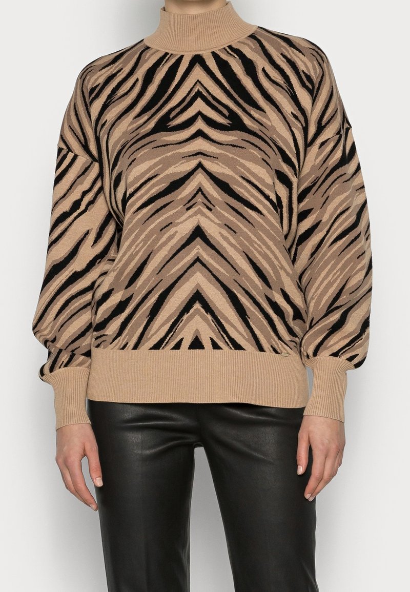 Pull à col roulé beige et noir à rayures tigre avec manches bouffantes, associé à un pantalon en cuir noir sur une personne debout.
