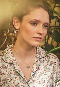 Ung kvinna med ljus hy och fräknar som bär en blommig blus, halsband med trollslända och matchande trollsländörhängen, med växter i bakgrunden.