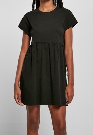 Kobieta w krótkiej czarnej sukience typu babydoll z podwiniętymi krótkimi rękawami i marszczoną talią, stojąca na jednolitym szarym tle.