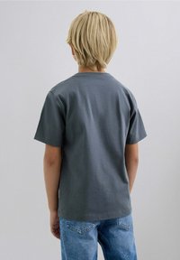 Camiseta de algodón gris con mangas cortas, escote redondo y un corte holgado, combinada con jeans de mezclilla azules. La vista es desde la parte de atrás.