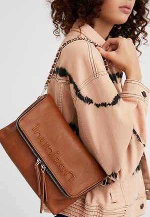 Frau trägt eine beige-schwarze Batikjacke und hält eine braune Lederhandtasche mit Kettenträger und eingeprägtem Markennamen auf der Klappe.