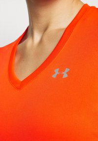 Top atletico a V in tessuto arancione brillante con una texture liscia. Presenta un piccolo logo grigio Under Armour sulla parte sinistra del petto.
