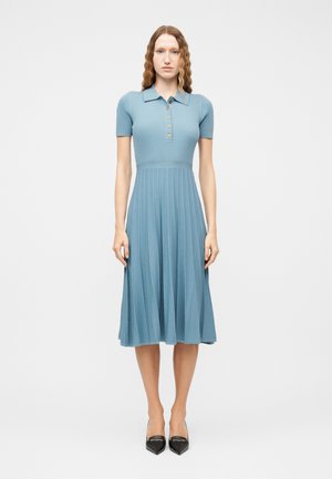 Kvinne som står vendt fremover iført en blå ribbestrikket kjole med korte ermer, knapper og plissert skjørt, svarte spisse hæler.