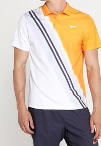 Homme portant un polo Nike à manches courtes bicolore orange et blanc avec des rayures noires diagonales et un short bleu marine.