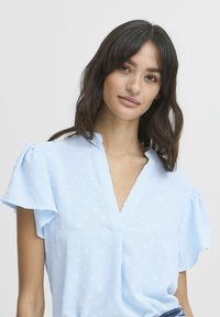 Blusa azzurra chiara con maniche corte e accenti arricciati, tessuto testurizzato e scollatura a V. Motivo a pois sottile su tutta la superficie.