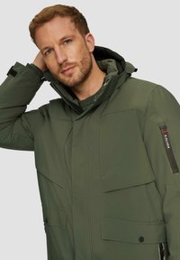 Grüne isolierte Jacke mit hohem Kragen und verstellbarer Kapuze. Verfügt über mehrere Taschen und einen Reißverschluss, hergestellt aus strapazierfähigem, glattem Stoff.