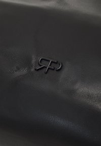 Produit en cuir noir présentant une surface texturée et un logo discrètement embossé. Le design est lisse avec des plis et des détails visibles.