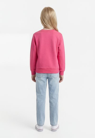 Laps pikkade blondide juustega, kandes roosat dressipluusi, helesiniseid teksaseid ja valgeid tossusid, seisab seljaga valgel taustal.