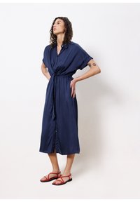 Marineblaues Hemdkleid mit kurzen Ärmeln, Taillengürtel und fließendem Schnitt. Kombiniert mit roten Riemchensandalen. Glatte Stoffoberfläche.