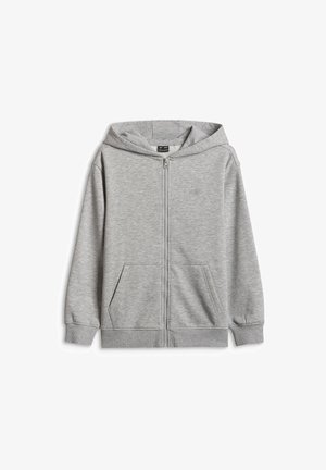 Garçon portant un bonnet bleu, un polo blanc, un sweat à capuche gris zippé, un pantalon de jogging gris et des baskets noires et blanches, debout les mains dans les poches.