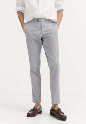 Uomo che indossa pantaloni slim fit grigio chiaro con le mani nelle tasche e mocassini in pelle marrone con nappine su sfondo bianco semplice.