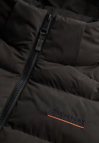 Gros plan sur une veste matelassée noire avec une fermeture éclair noire et le logo "Superdry Sportswear" brodé près de l'extrémité de la fermeture éclair.