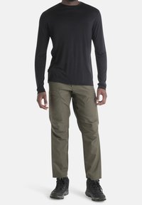 Mann in einem schwarzen Langarmshirt, olivgrünen Cargo-Hosen und schwarzen Wanderschuhen vor einem einfarbigen Hintergrund stehend.